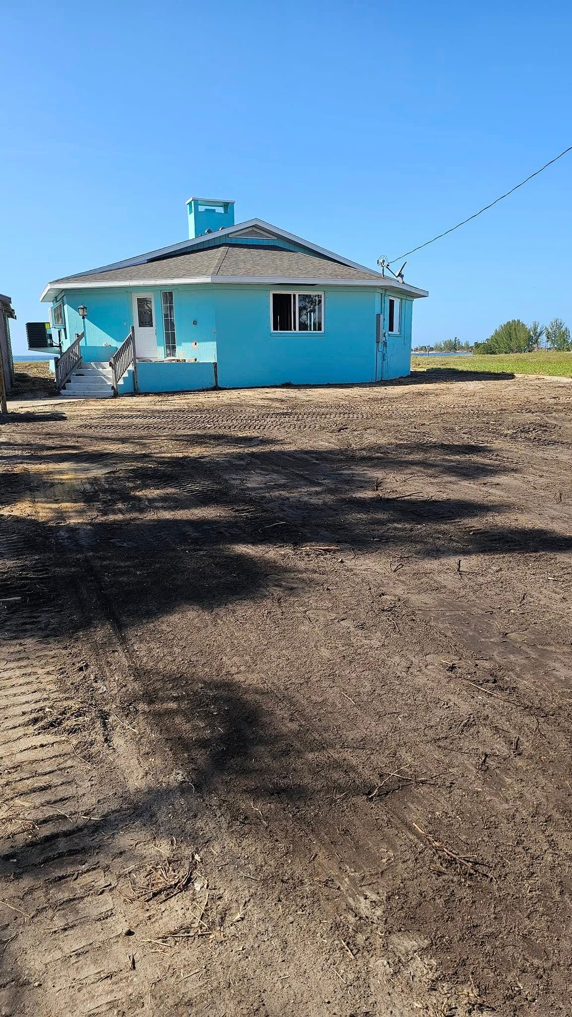 A bright blue, round house with a gray roof sits on a large, empty dirt lot under a clear blue sky.