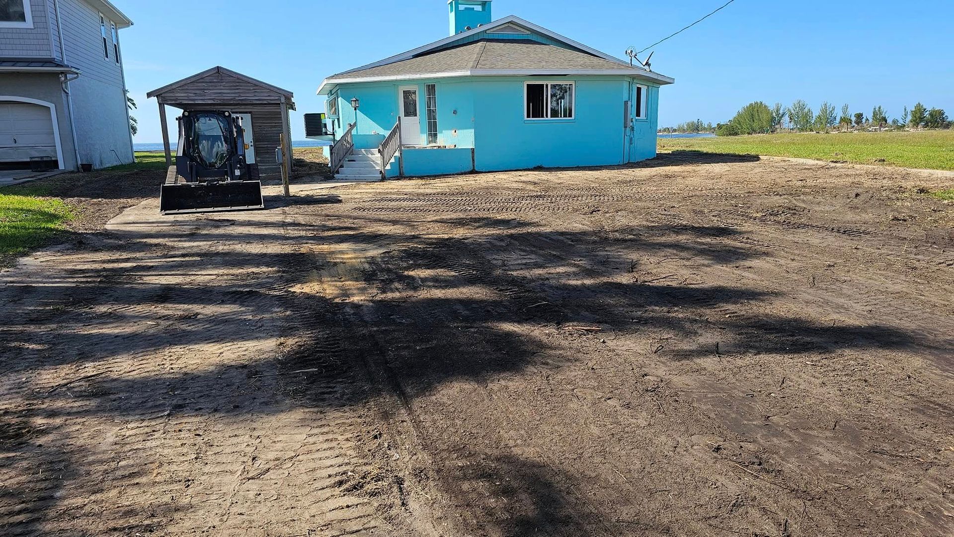 A construction skid-steer loader sits in a dirt lot next to a bright blue house on a sunny day.