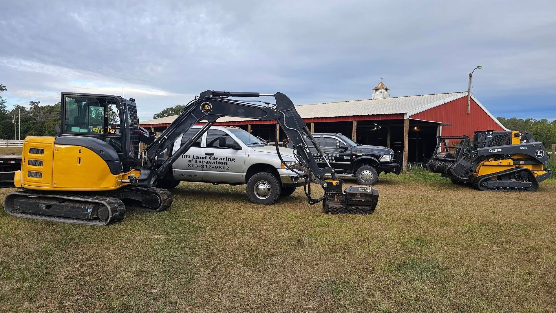 A yellow excavator, a silver pickup truck, and a tracked loader parked in a grassy field in front of a red barn.