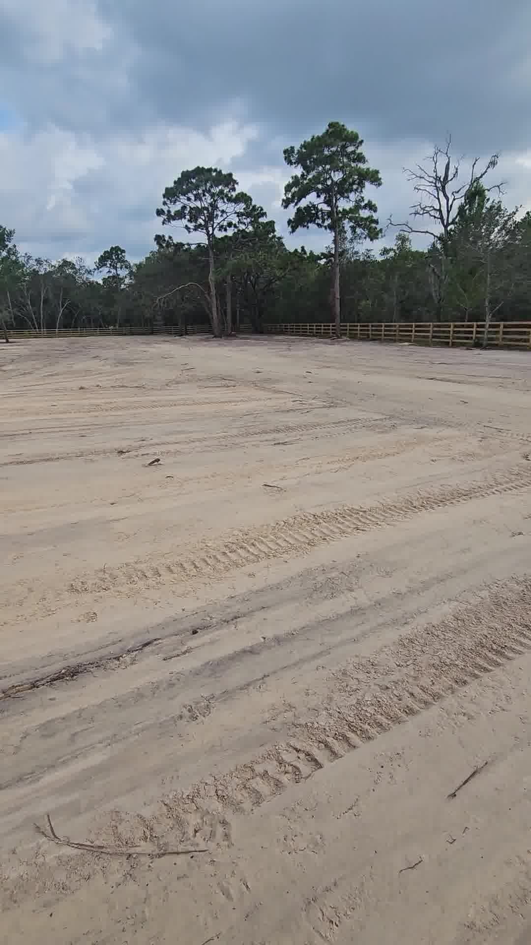 Wide, flat, sandy lot showing tire tracks leading toward a distant wooden fence and trees under a cloudy sky.