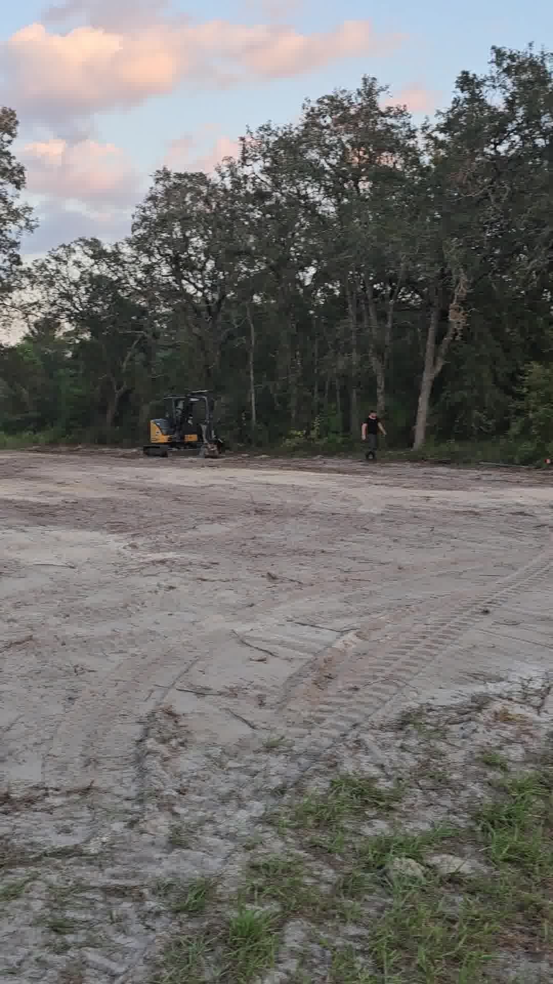 A person stands near a small yellow excavator on a cleared, sandy construction site bordered by a forest at sunset.