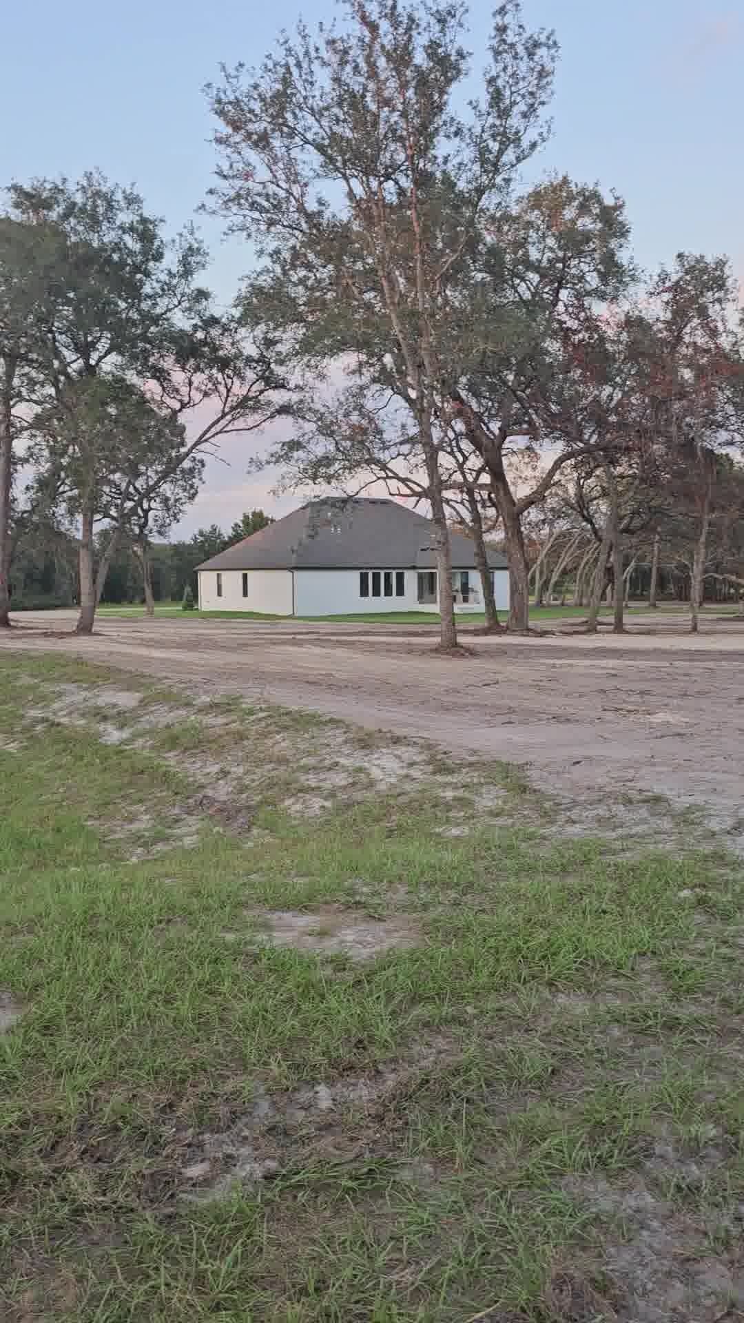 A white, single-story house stands in a large, sandy clearing surrounded by mature trees under a pale sunset sky.