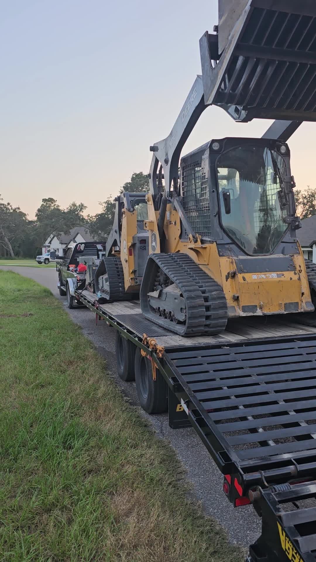 A yellow compact track loader secured on a flatbed trailer parked on a road beside a grassy area.