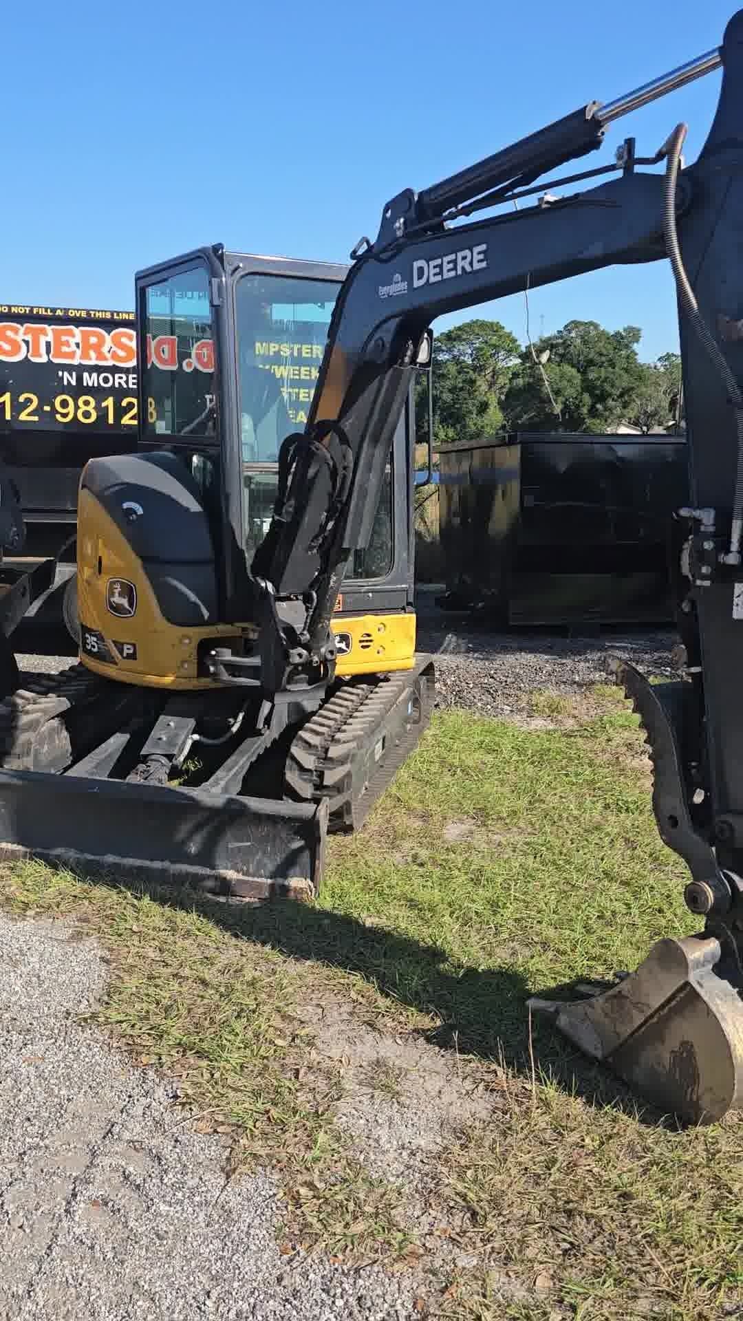 A yellow and black John Deere compact excavator parked on a gravel lot near a dumpster under a clear blue sky.