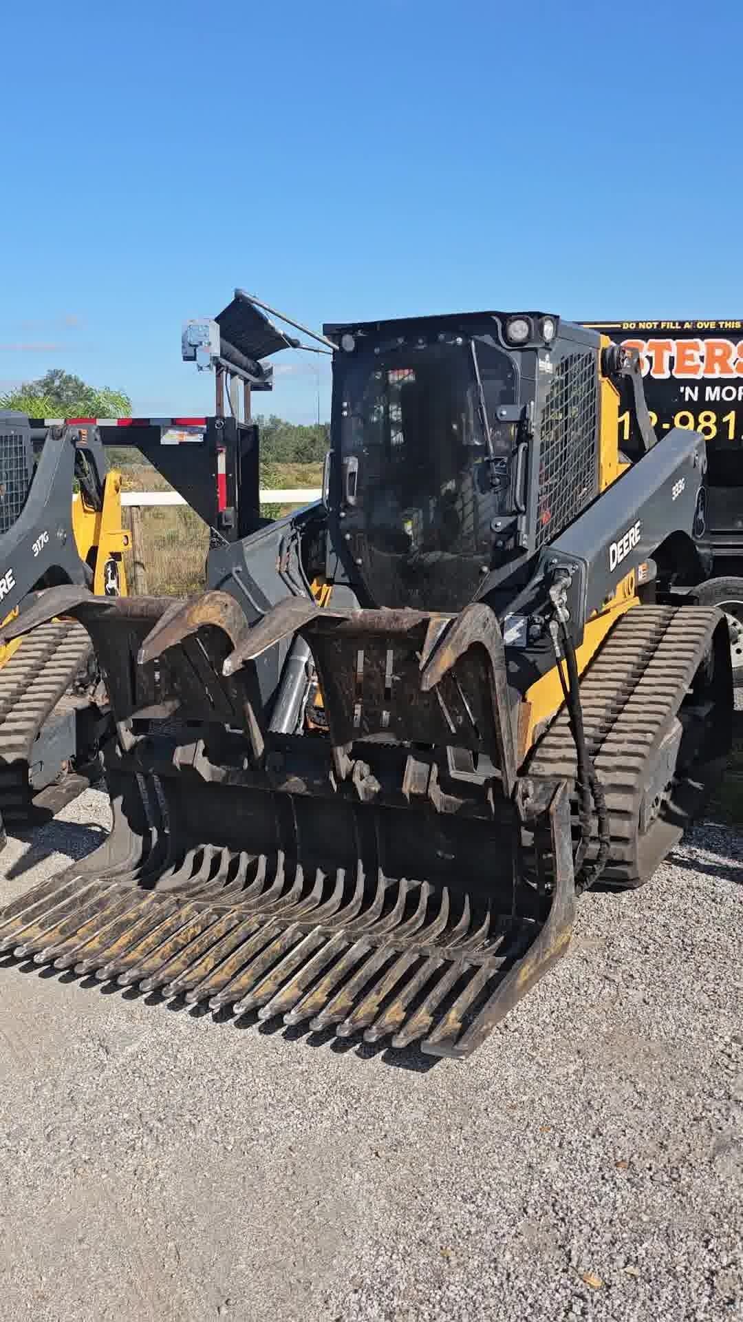 Yellow and black compact track loader with a metal grapple bucket attachment parked on a gravel lot under a blue sky.