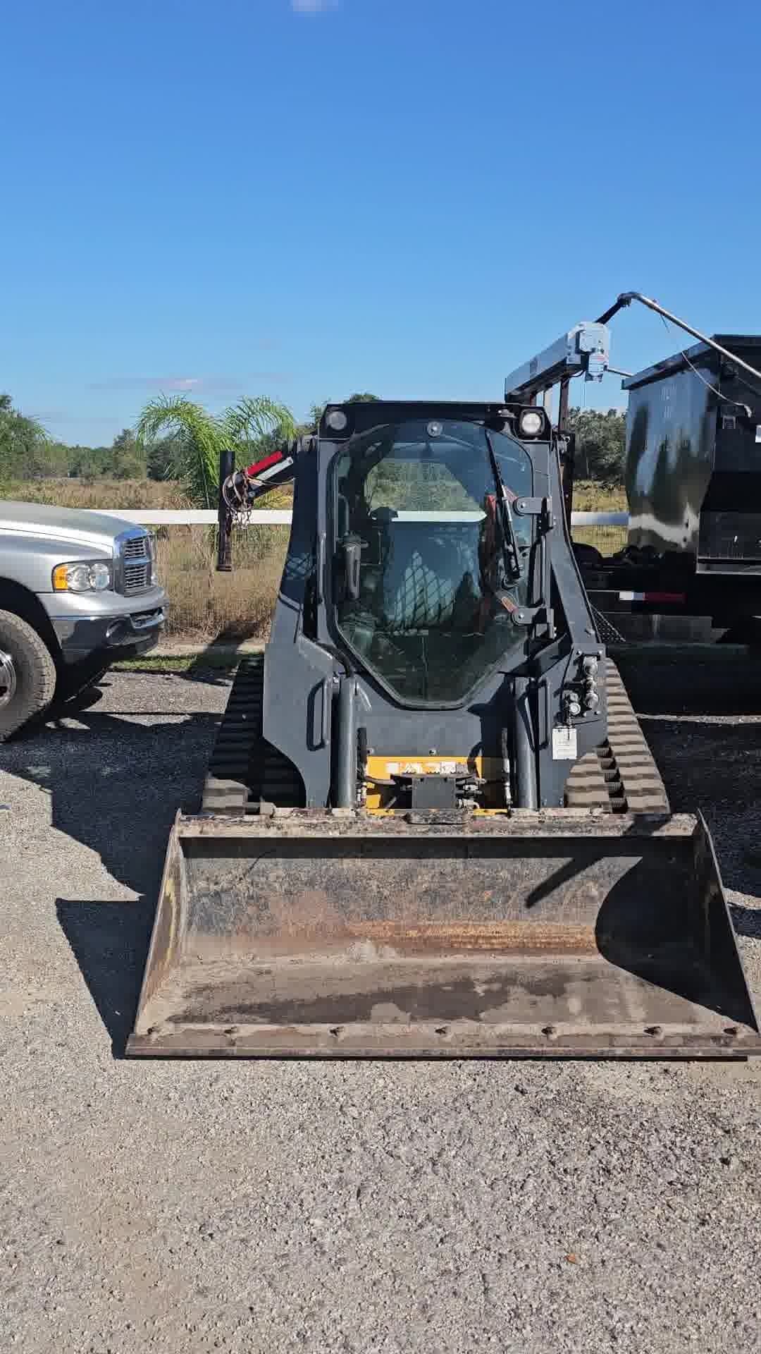A dark gray compact track loader with a large bucket attachment parked on a gravel lot next to a pickup truck and trailer.