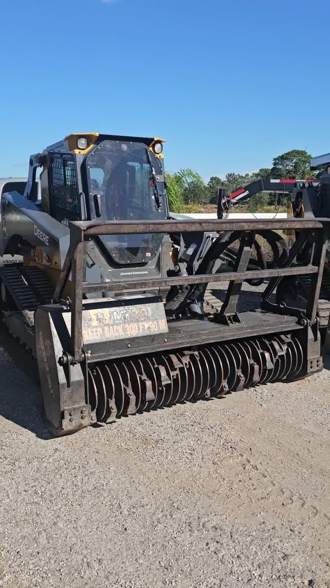 A dark-colored skid steer loader with a large forestry mulcher attachment parked on a gravel lot under a clear blue sky.
