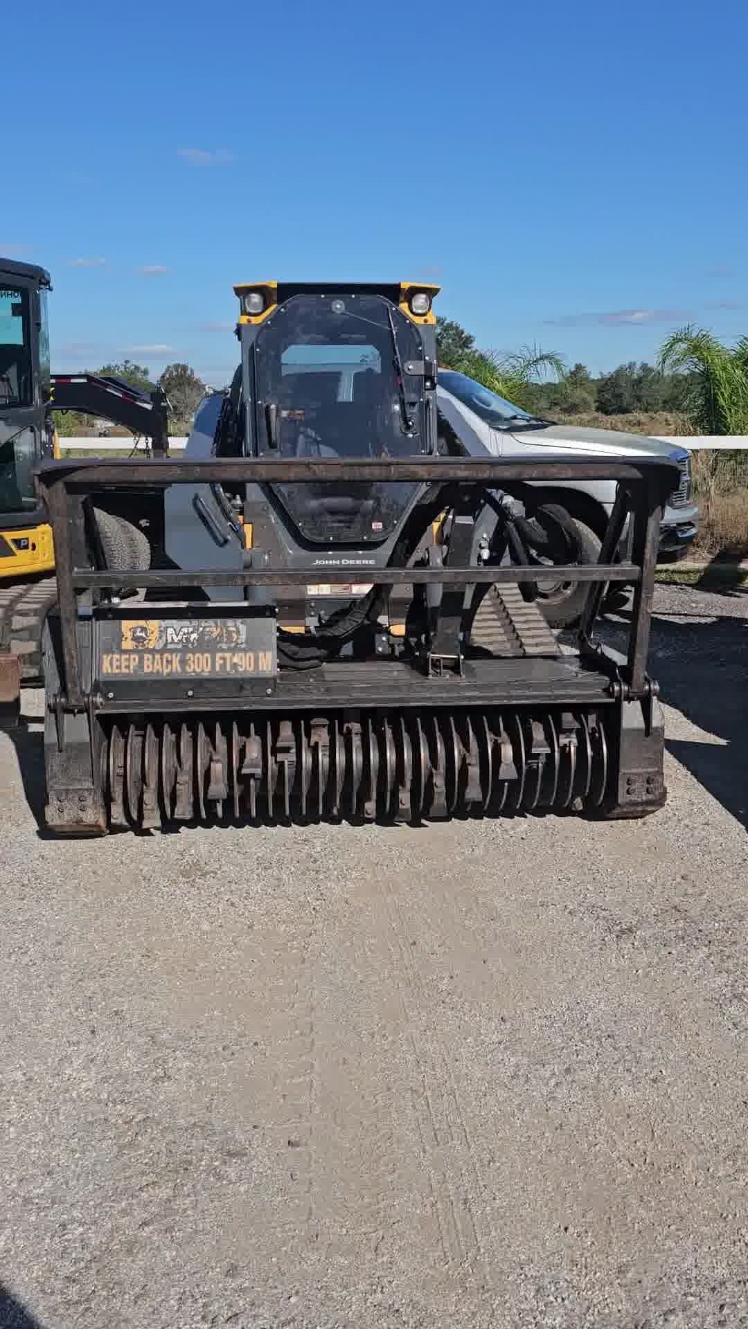 A skid-steer loader equipped with a land-clearing forestry mulcher attachment, parked on a gravel lot under a clear sky.