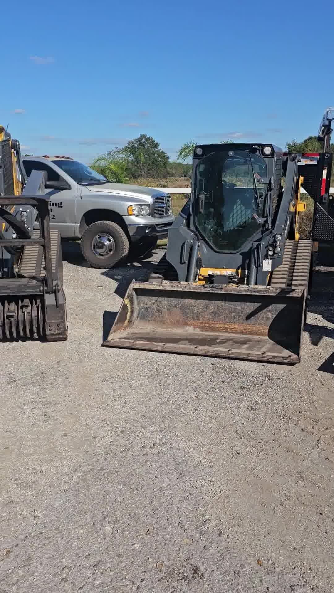 A skid steer loader sits on a gravel lot next to a parked gray pickup truck under a clear blue sky.