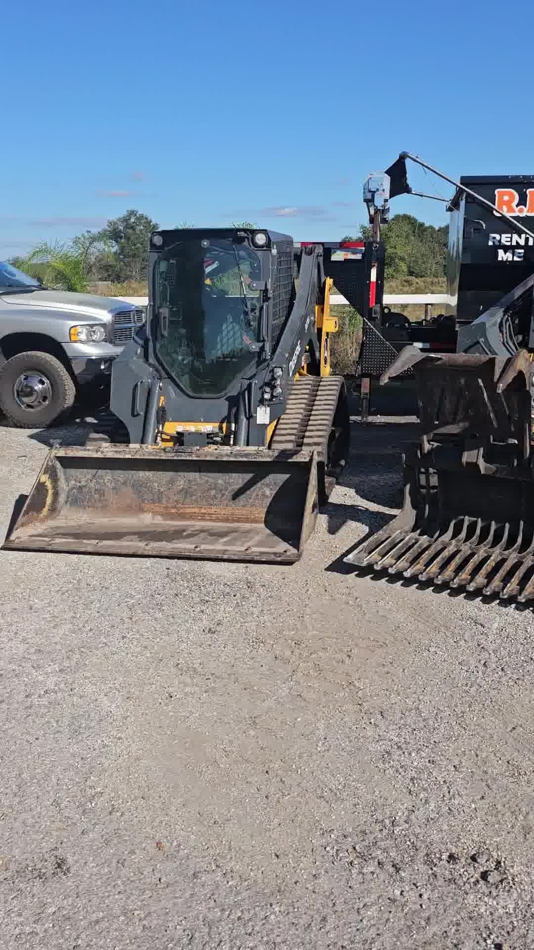 A dark grey compact track loader parked on a gravel lot next to a pickup truck and a separate metal rake attachment.
