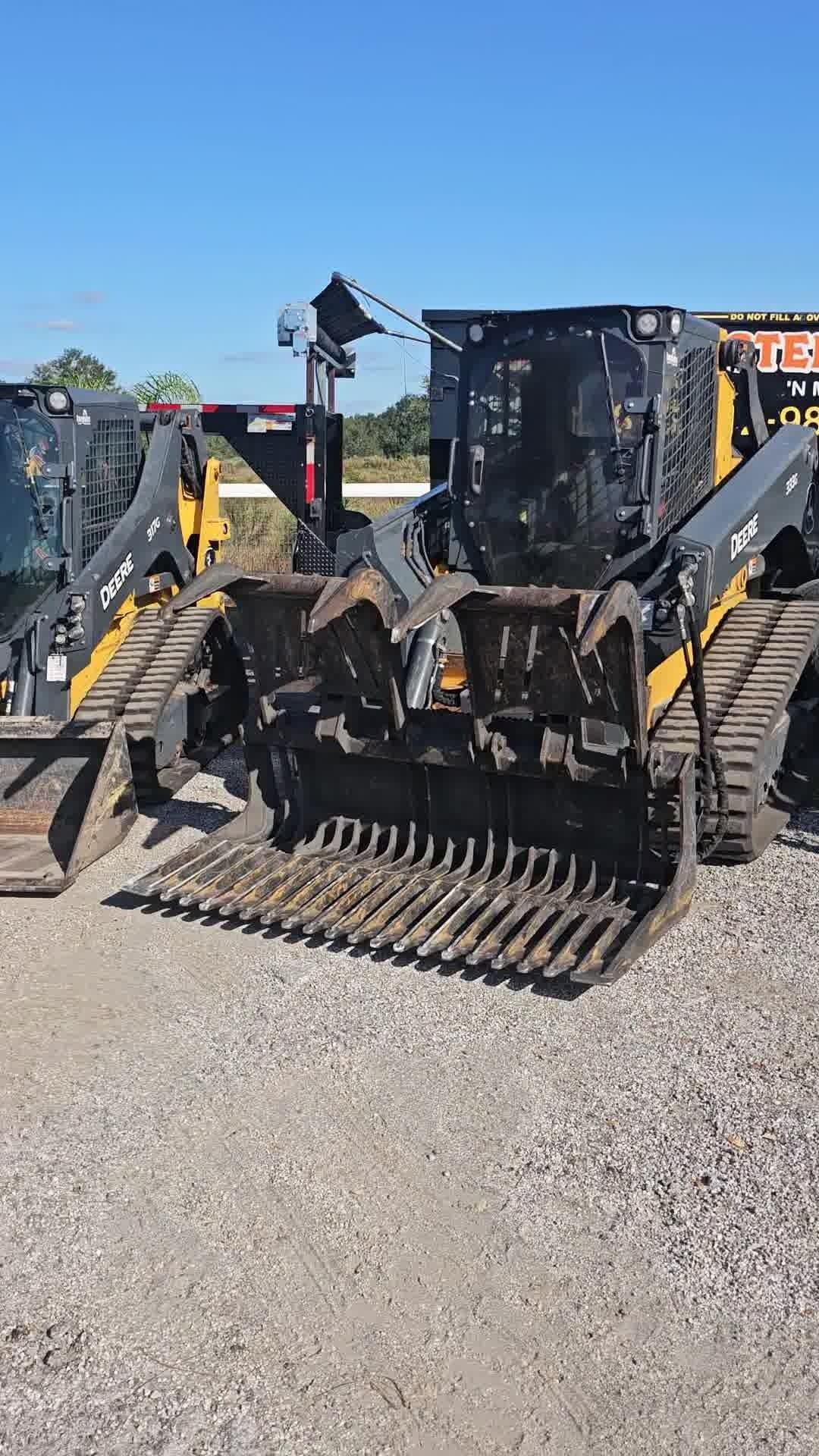 Two yellow and black tracked skid steer loaders with skeleton rock buckets parked on a gravel lot under a clear blue sky.