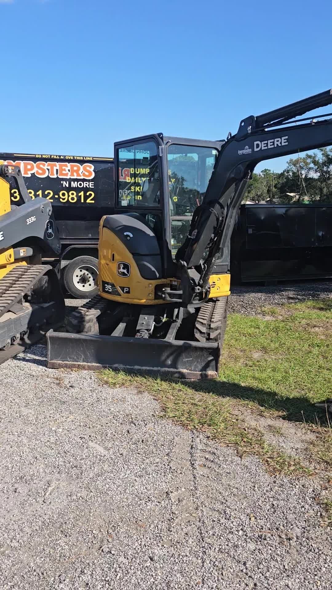 A yellow and black John Deere compact excavator parked on a gravel lot under a bright blue sky.