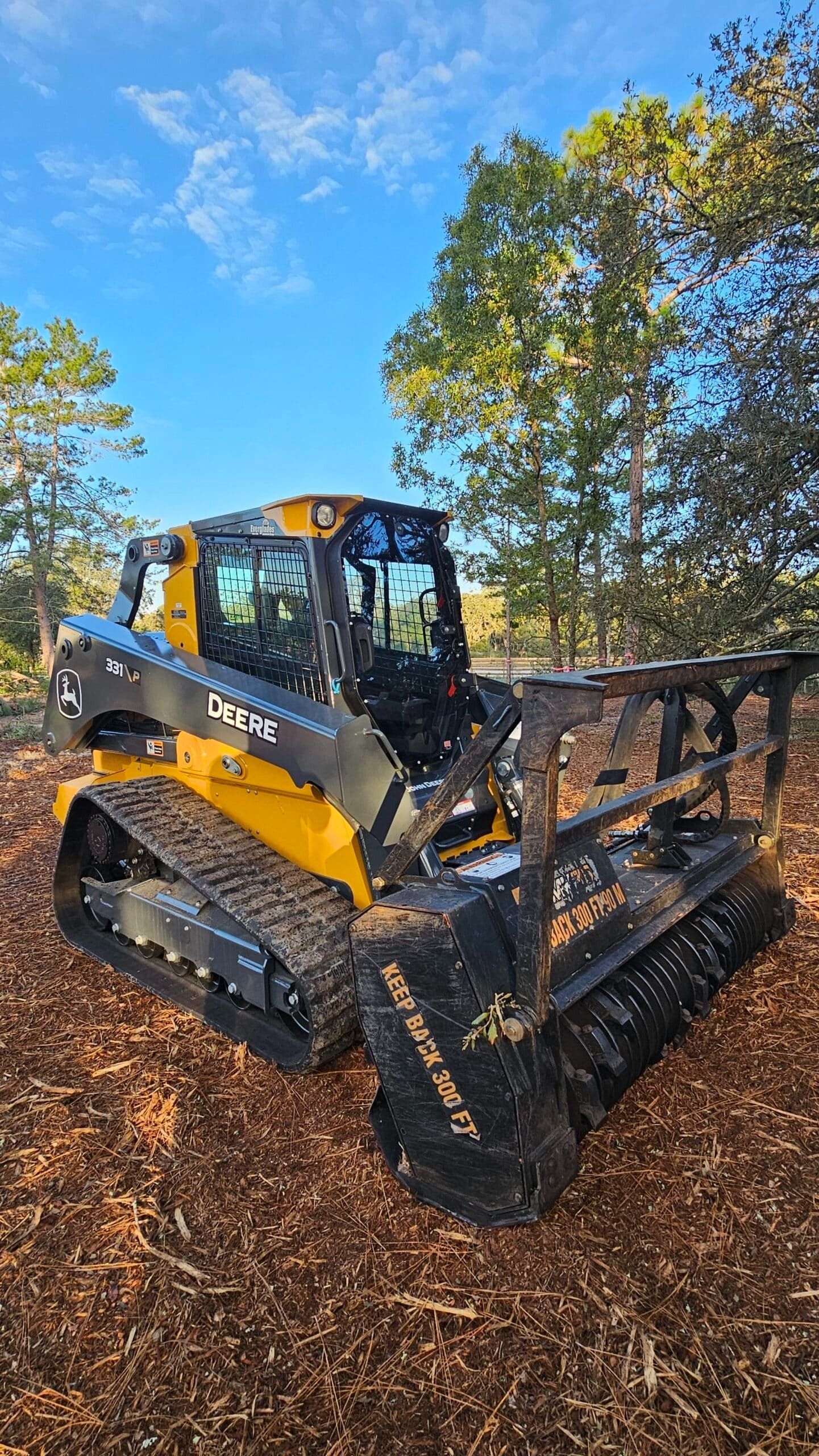 A yellow and black John Deere compact track loader with a forestry mulcher attachment sits in a wooded area.