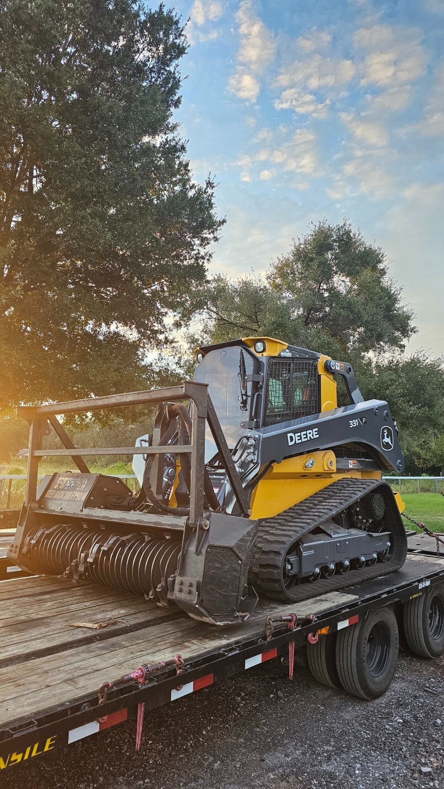 Yellow and black track loader with a forestry mulcher attachment secured on a flatbed trailer outdoors at sunset.