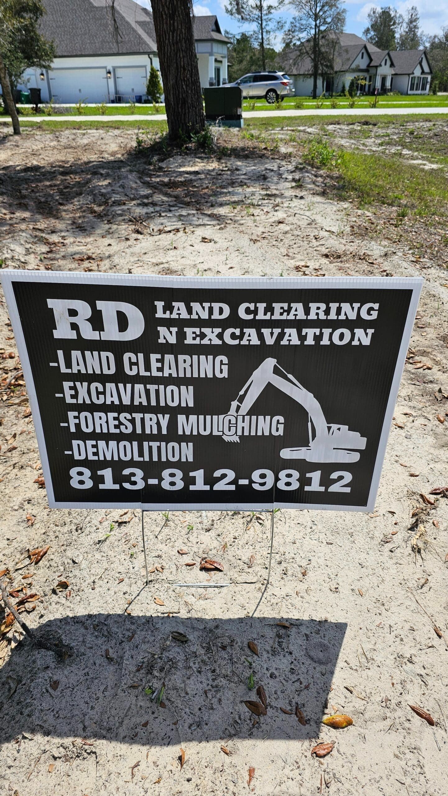A black and white sign for RD Land Clearing N Excavation, listing services and the phone number 813-812-9812 on a sandy lot.