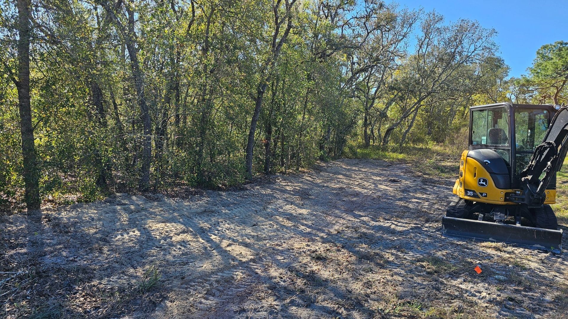 A yellow compact excavator parked on a cleared, sandy patch of land next to a dense line of trees on a sunny day.