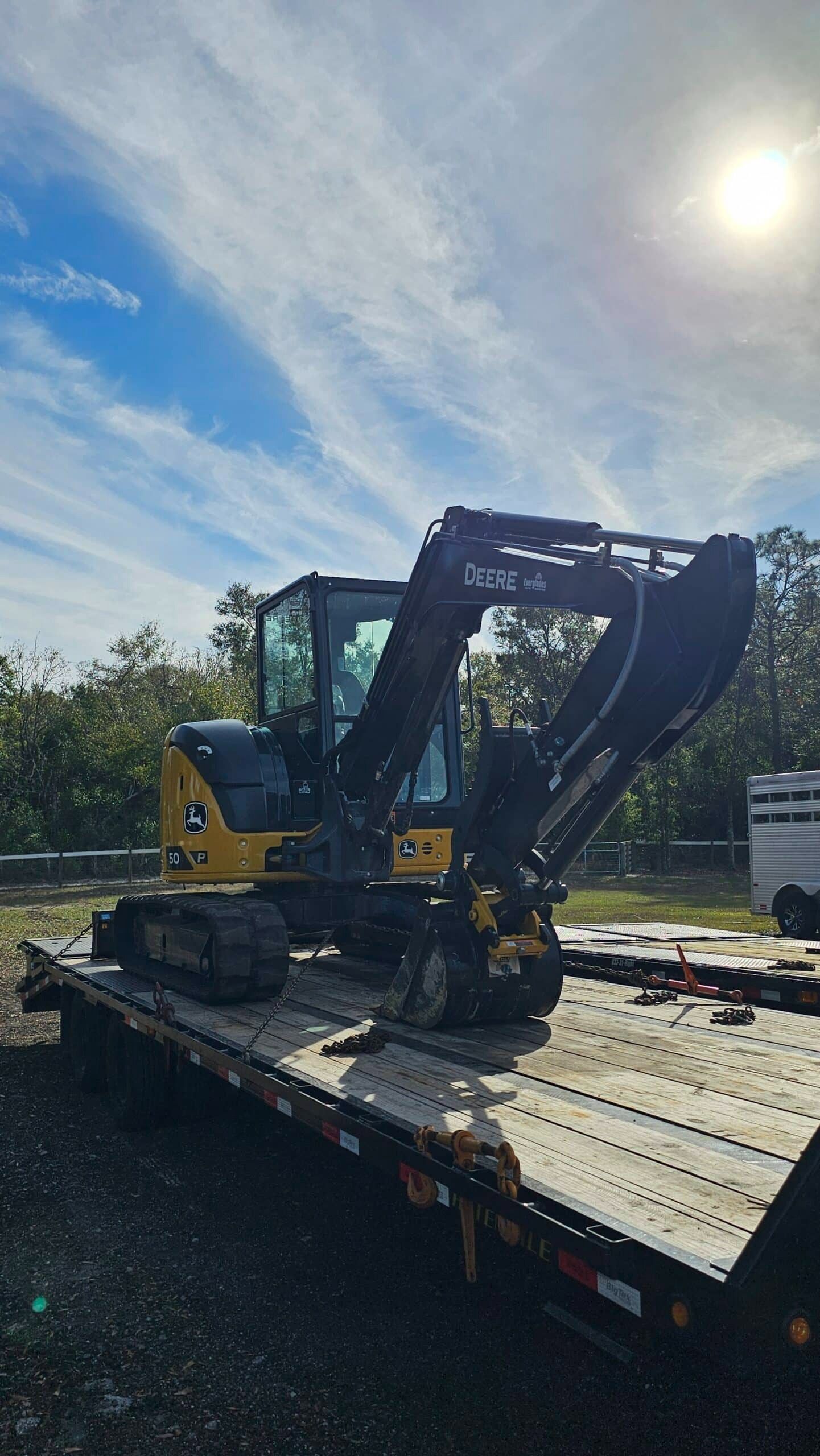 A yellow and black John Deere excavator loaded onto a flatbed trailer outdoors under a bright, sunny sky.