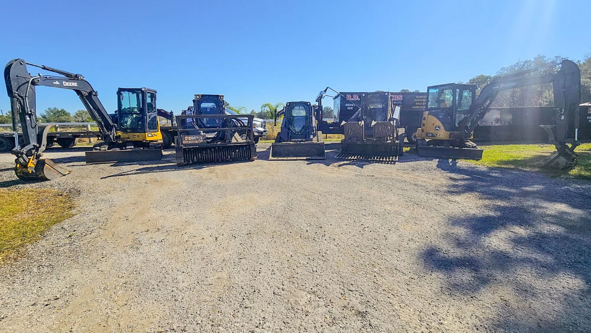 A line of yellow and black construction machinery, including excavators and skid steers, parked on a gravel lot.