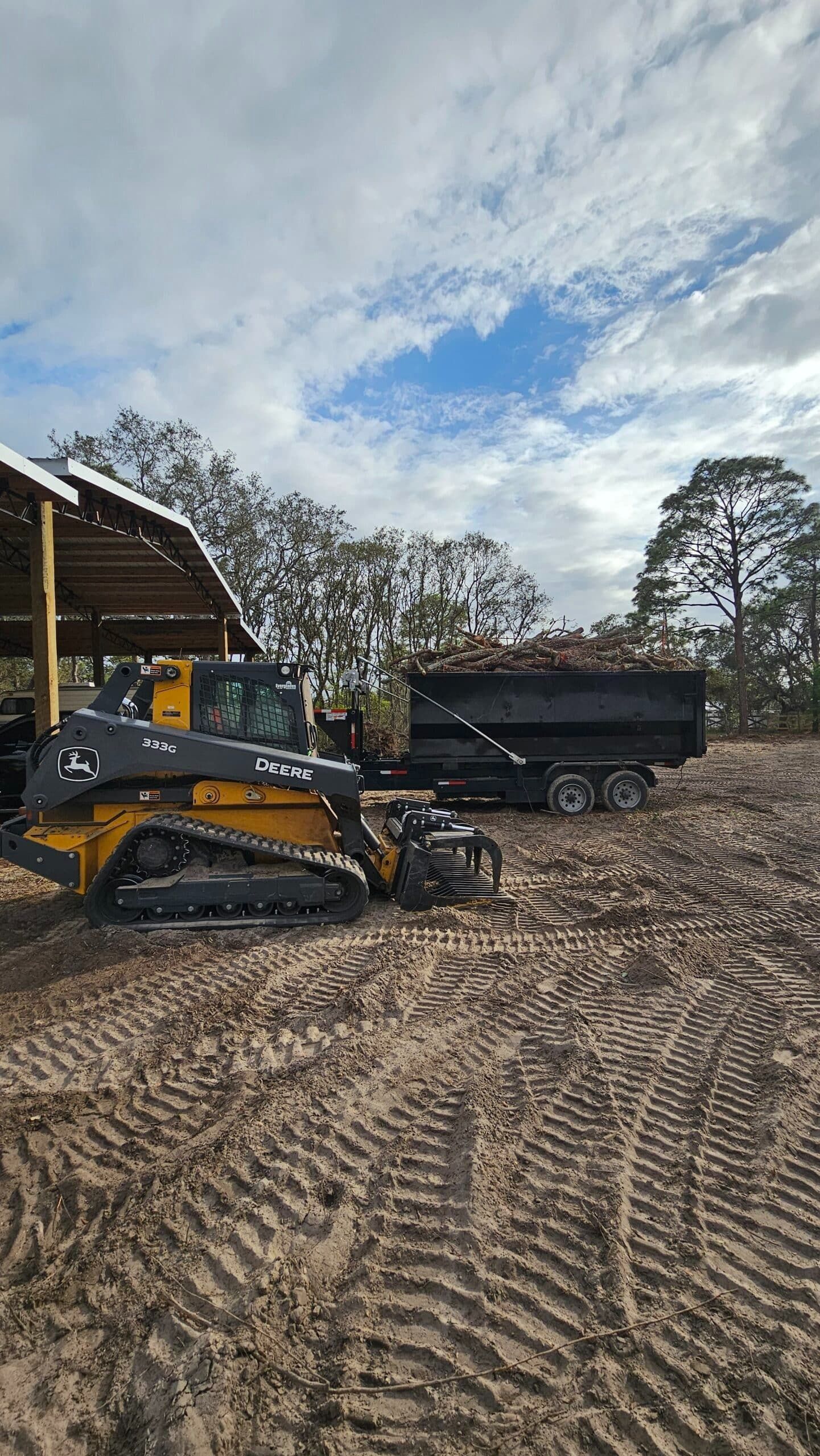 A yellow tracked skid steer parked next to a black dump trailer filled with debris at a cleared outdoor work site.