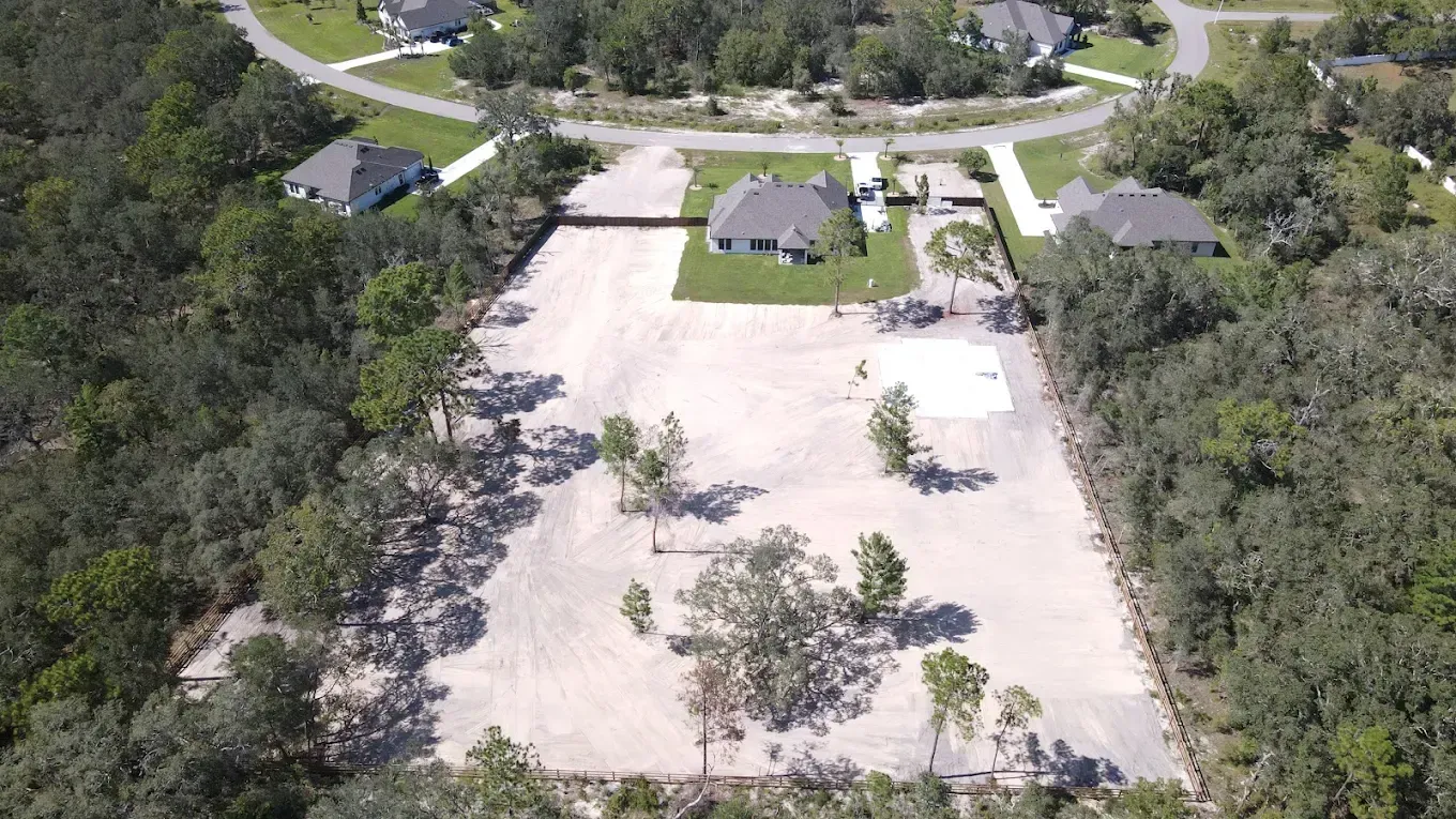 An aerial view of a large, cleared dirt lot situated among houses and surrounding woods.