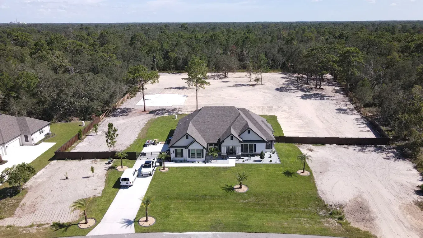 Aerial view of a white house with a gray roof, surrounded by a large gravel yard and dense forest.