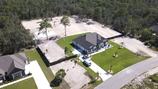 An aerial view of a single-story house with a black roof, a green lawn, and a large, cleared sandy lot behind it.