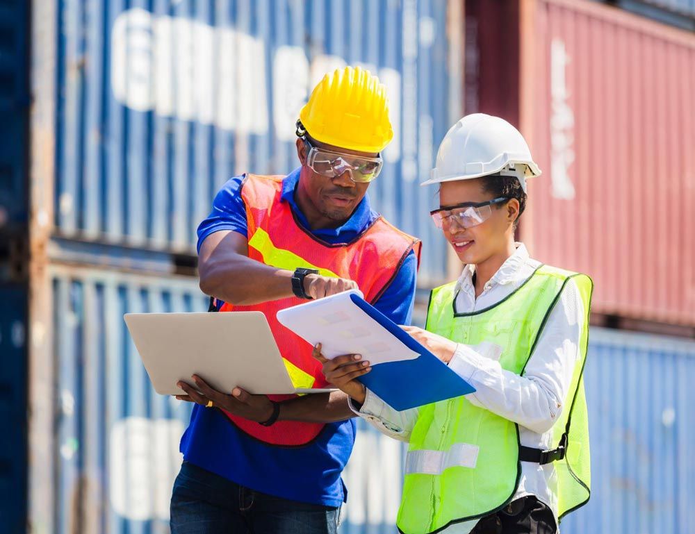 Two workers in safety gear review documents at a shipping port, with stacked containers in the background.
