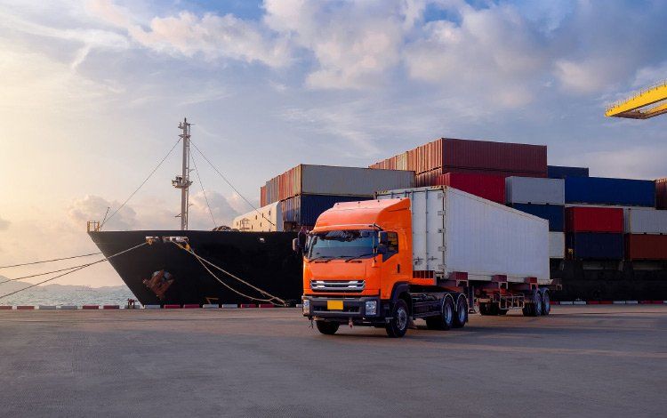 Orange truck on a dock beside a cargo ship with shipping containers under a cloudy sky.
