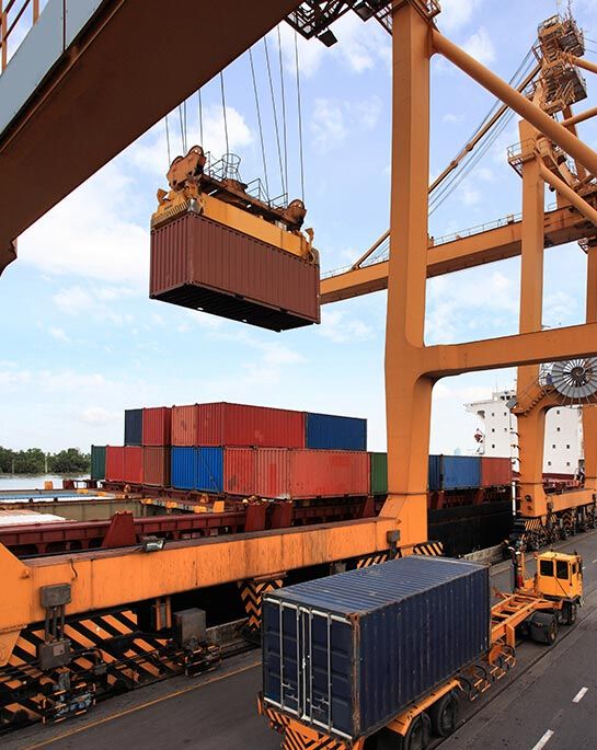 Container crane lifting a shipping container at a port, other containers and a semi-trailer nearby.