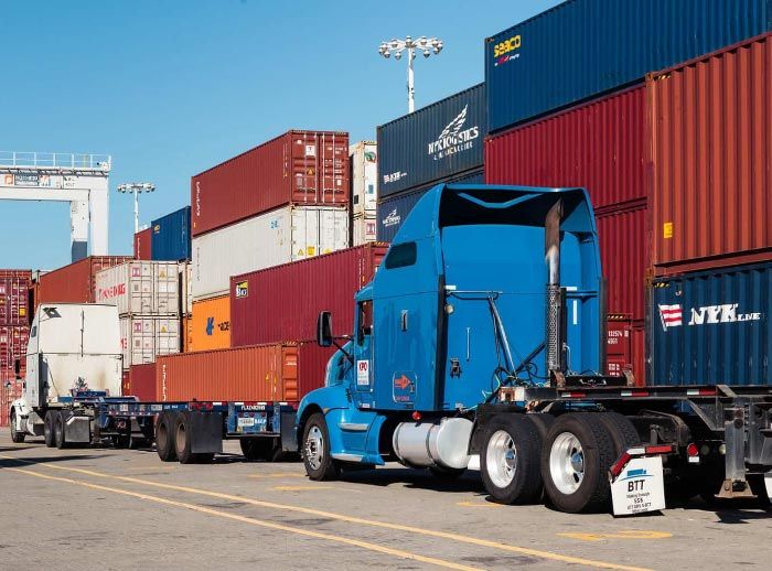 Blue semi-truck loaded with shipping containers at a port. Stacked containers are red and blue.