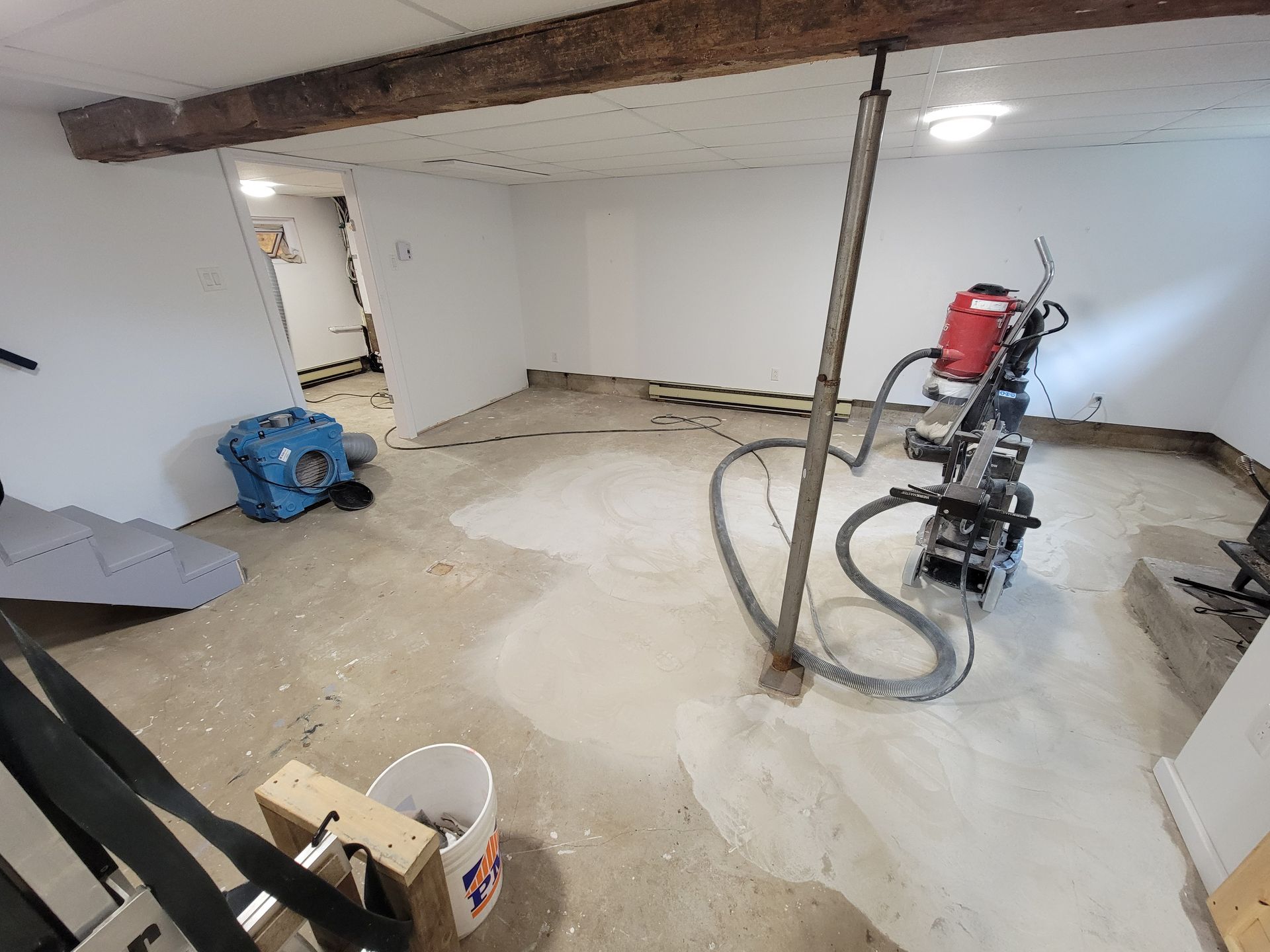 Sous-sol en rénovation; plancher de béton avec meuleuse, murs blancs, poteau de support, porte d'entrée.