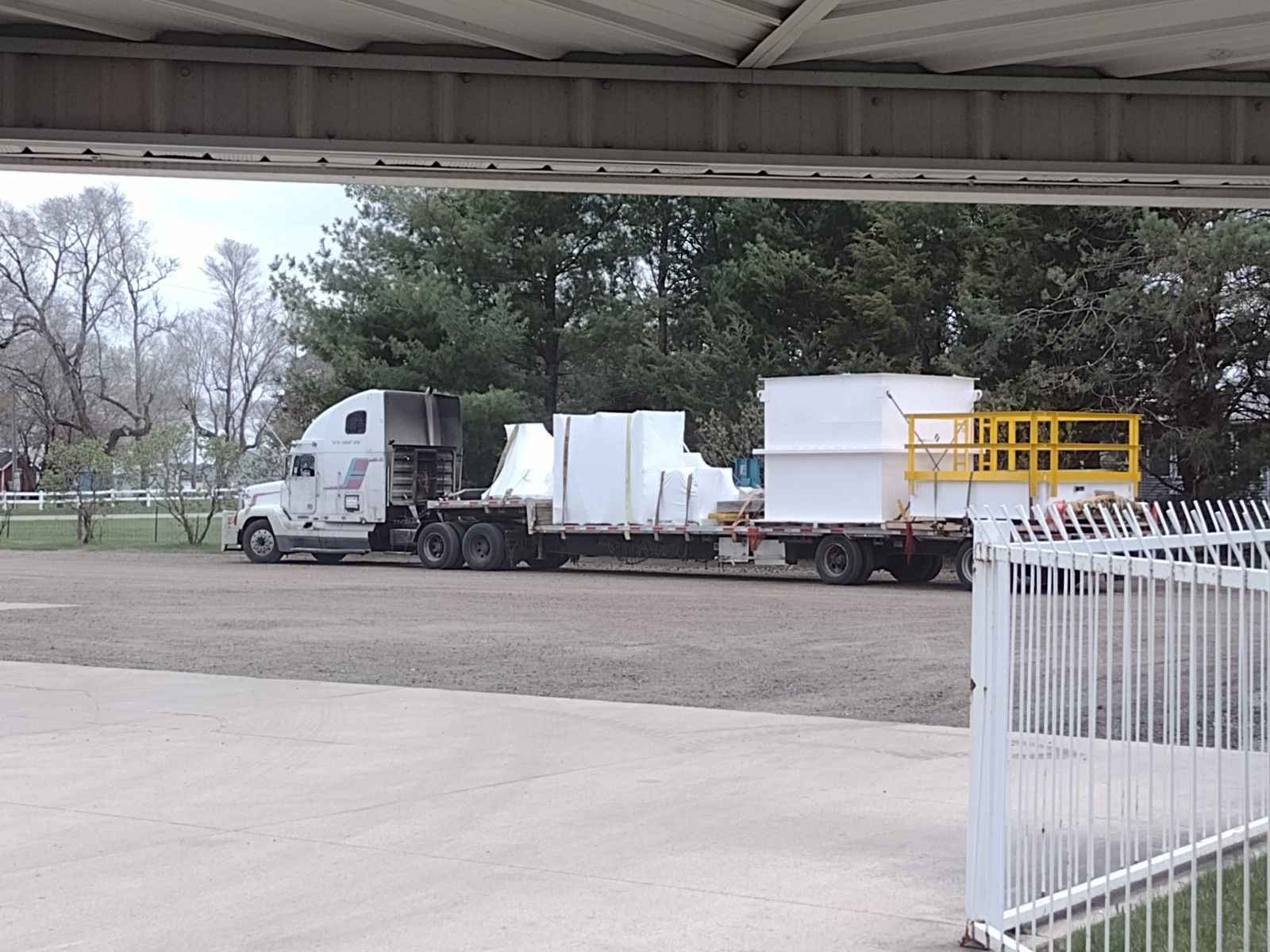 White semi-truck hauling equipment parked by a road under a bridge, with a white fence in the foreground