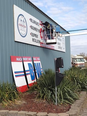 Construction sign on blue metal building, with workers on a platform near a wall-mounted sign and tall plants below.