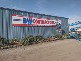 B&W Contractors sign on blue metal building with a worker on blue equipment beside a dirt road