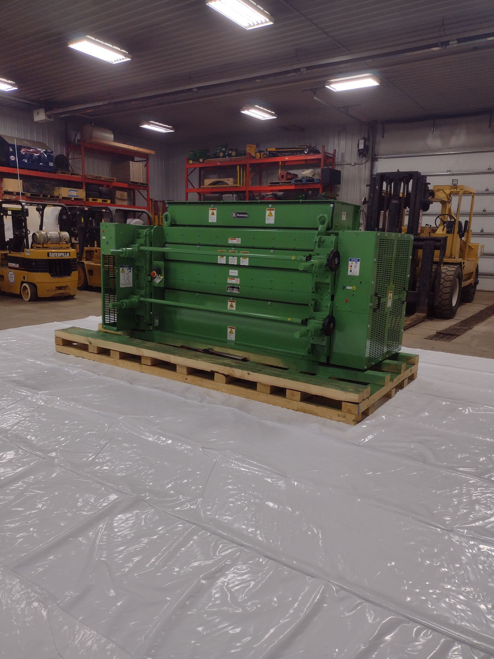 Green industrial machine equipment on a wooden pallet in a warehouse, with forklifts in the background
