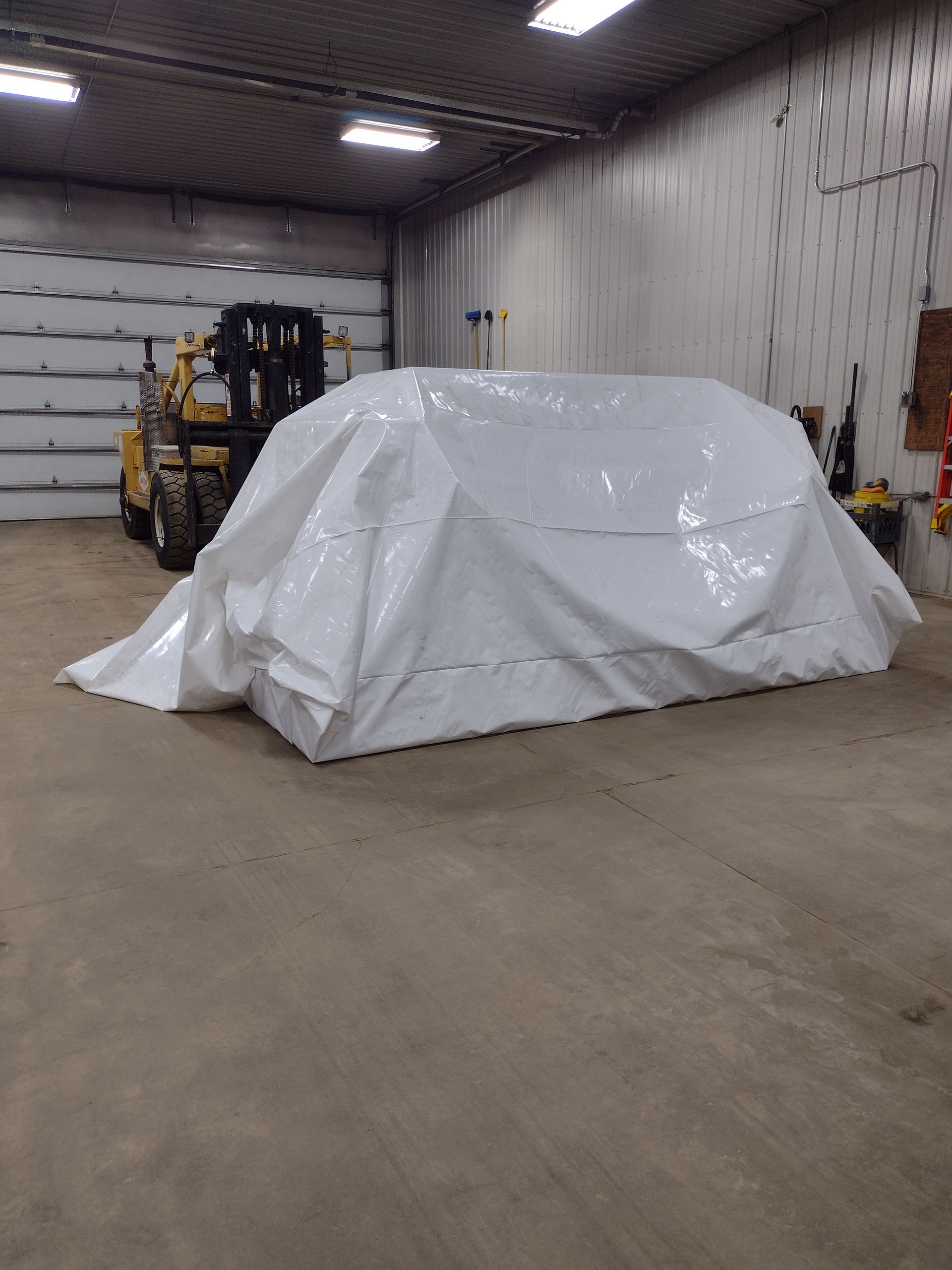White tarp-covered object in a warehouse beside a forklift and garage door.