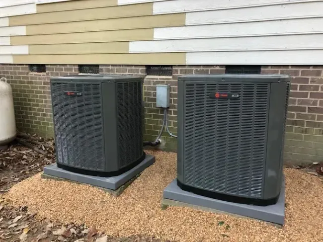 Two dark-colored air conditioning units on concrete pads next to a building with brick and siding, on a bed of gravel.