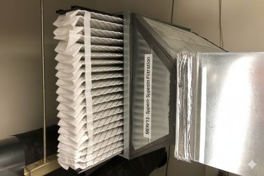 Air filter within metal ductwork, side view. Pleated white filter, labeled 
