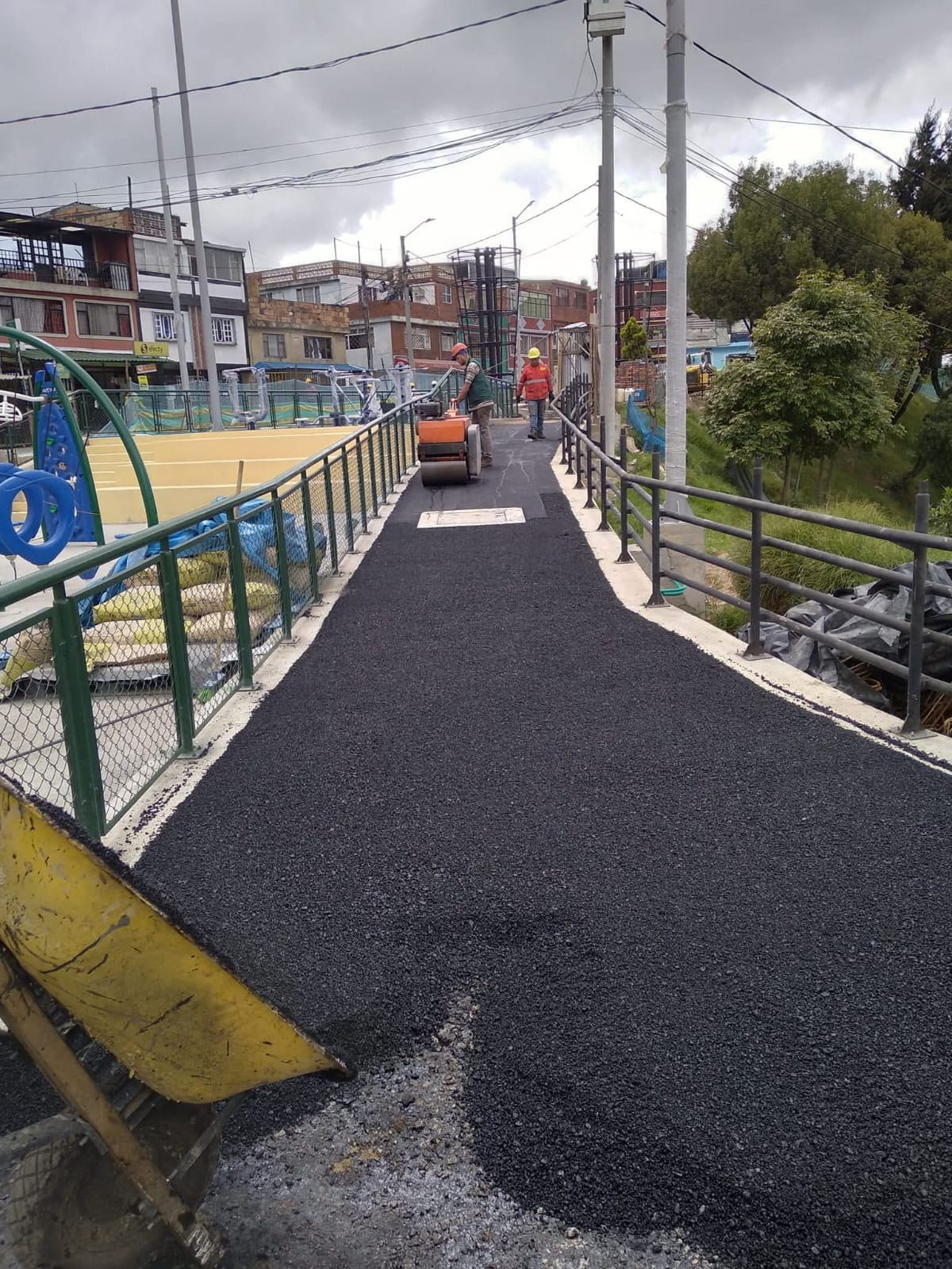 Puente peatonal estrecho con pavimento negro y barandillas verdes, que conduce hacia unos edificios en una zona urbana nublada