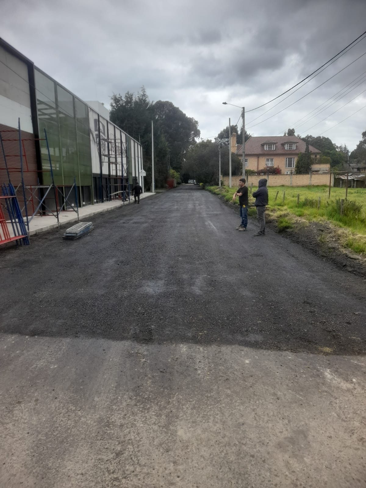 Camino de grava junto a un edificio de cristal, con dos personas cerca de un campo de hierba bajo un cielo nublado.