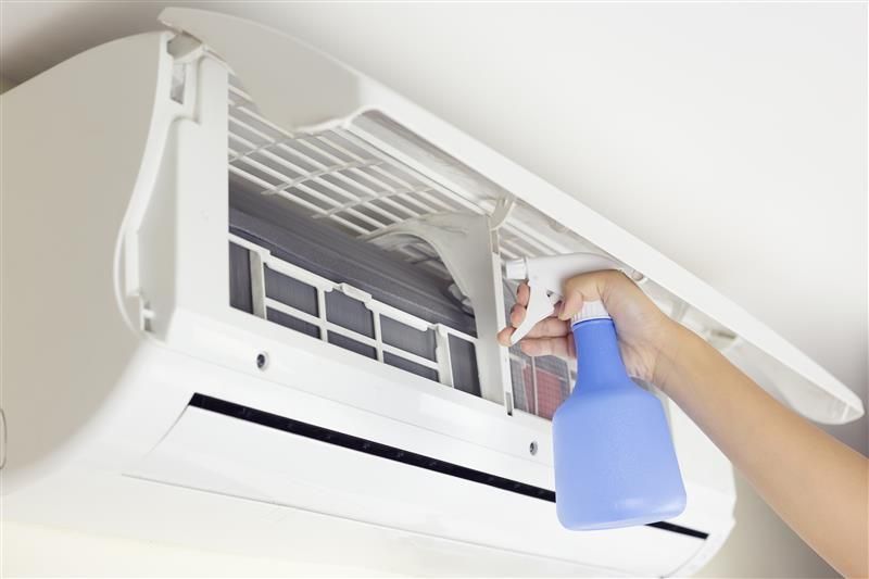 Person sprays an air conditioner's filter with a blue bottle, inside a white air conditioning unit.