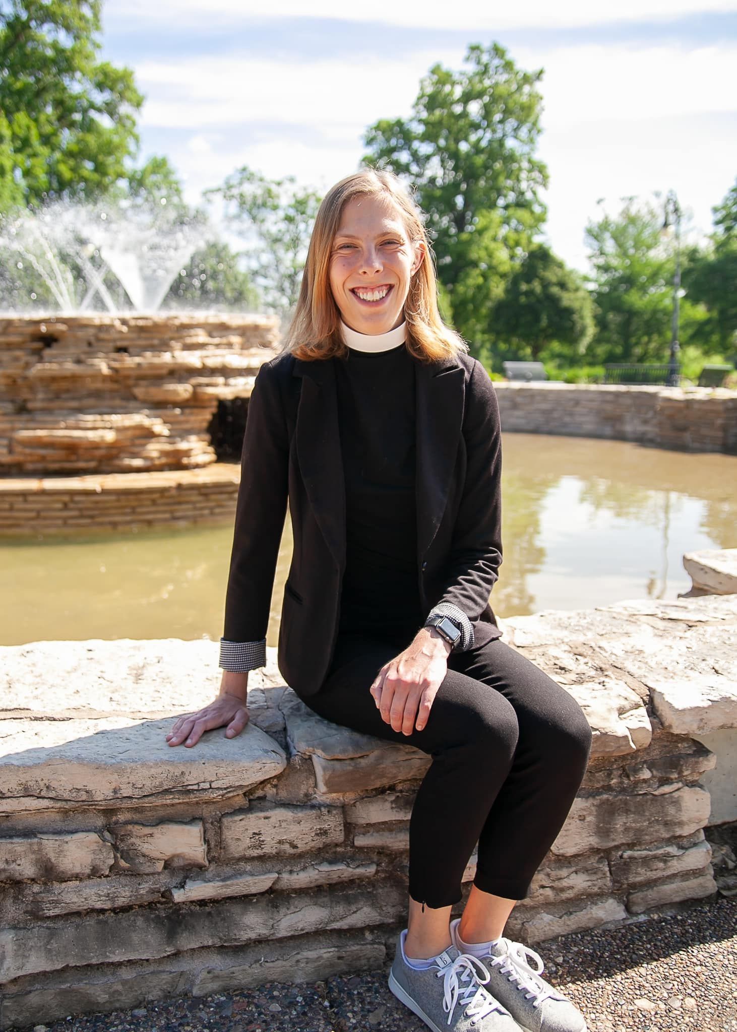 A woman is sitting on a stone wall in front of a fountain.