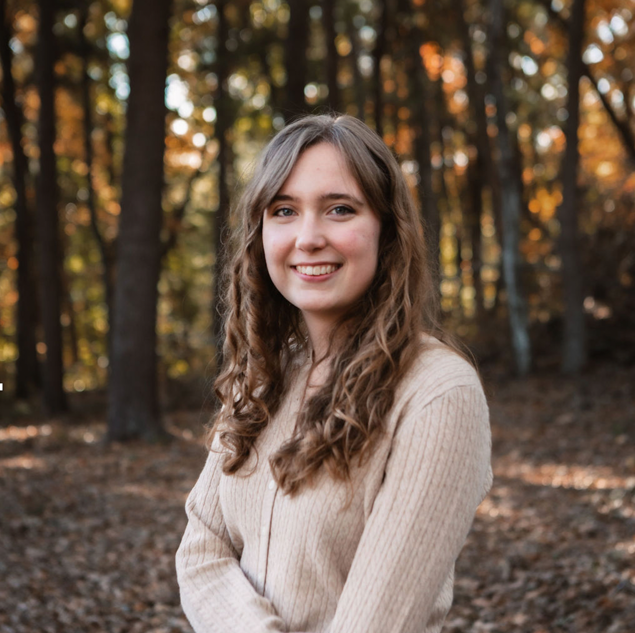 A woman in a tan sweater is standing in the woods.
