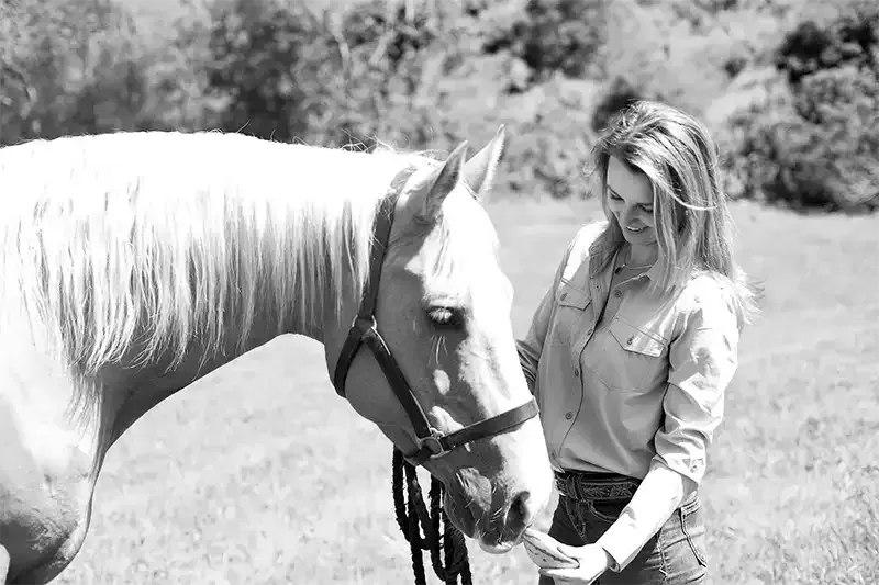 Woman smiles, gently touching the muzzle of a palomino horse in a grassy field.