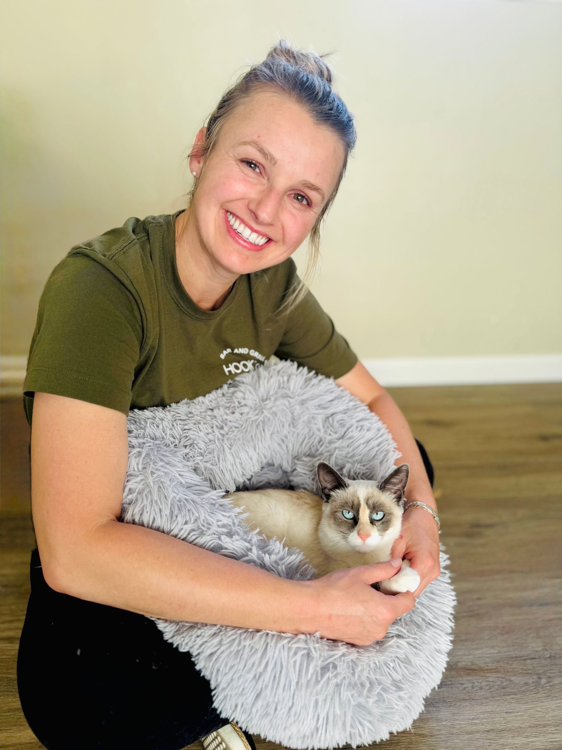 Woman smiling while holding a cat in a gray bed on a hardwood floor