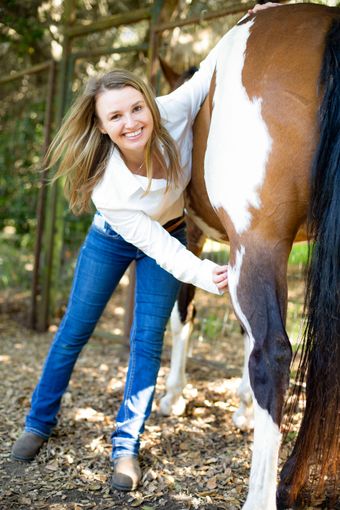 Woman smiling beside a brown-and-white horse in a wooded area