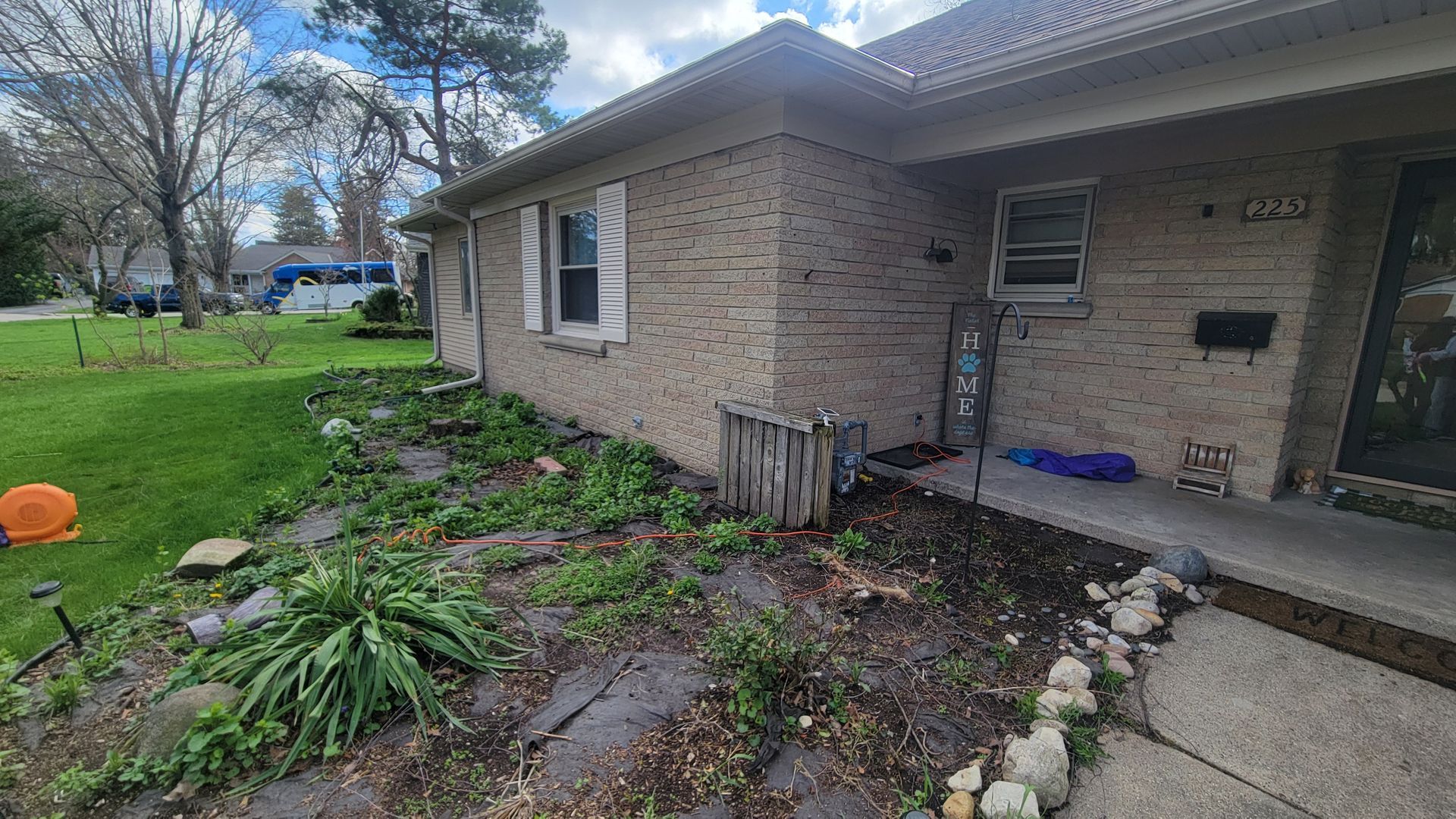 Brick house corner with a front porch, concrete walkway, and a small garden bed in the yard.
