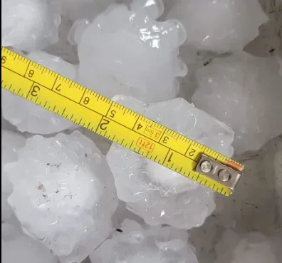 A yellow tape measure positioned over a cluster of large, jagged hailstones, showing a diameter of about 2 inches.