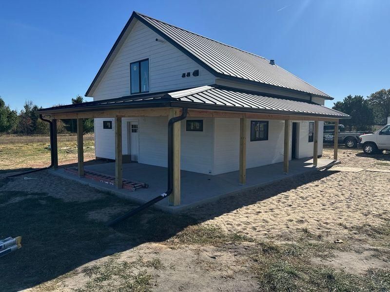 A white house with a metal roof and a wooden wrap-around porch stands on a dirt lot.