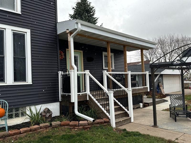 A dark-sided house with a small covered porch, white railing, stairs, and a white downspout.